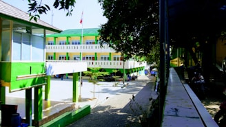 A modern, three-story building with a green and white color scheme is situated within a campus or school environment. The architecture features clean lines and a shaded walkway with trees and a flagpole in the courtyard. Motorcycles are parked along a pathway, indicating it might be a busy area.