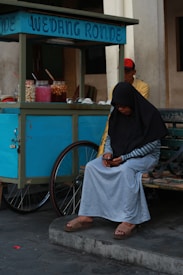 A person wearing a black hijab and gray skirt is sitting on a bench beside a street stall. The stall is labeled 'Wedang Ronde' and has jars of colorful ingredients. A man in a yellow shirt and red cap is partially visible behind the cart. The setting appears to be a casual outdoor environment.