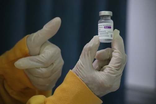 Gloved hands are holding a vial labeled 'Covid-19 Vaccine AstraZeneca'. One hand is giving a thumbs-up gesture, indicating approval or success.