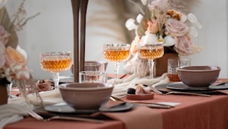 A beautifully set dining table with an elegant and soft aesthetic. Crystal glasses filled with amber-colored liquid are prominently displayed alongside ceramic bowls and plates on a terracotta-colored tablecloth. The table is adorned with lush floral arrangements featuring pastel roses and dried grasses, contributing to a sophisticated and calming ambiance.