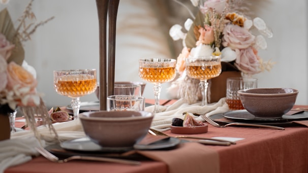 A beautifully set dining table with an elegant and soft aesthetic. Crystal glasses filled with amber-colored liquid are prominently displayed alongside ceramic bowls and plates on a terracotta-colored tablecloth. The table is adorned with lush floral arrangements featuring pastel roses and dried grasses, contributing to a sophisticated and calming ambiance.