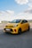 A bright yellow hatchback car is parked on a smooth pavement with a clear blue sky and fluffy white clouds above. The car features black alloy wheels and red accents on the front grill and bumper. In the background, there are distant buildings and greenery.