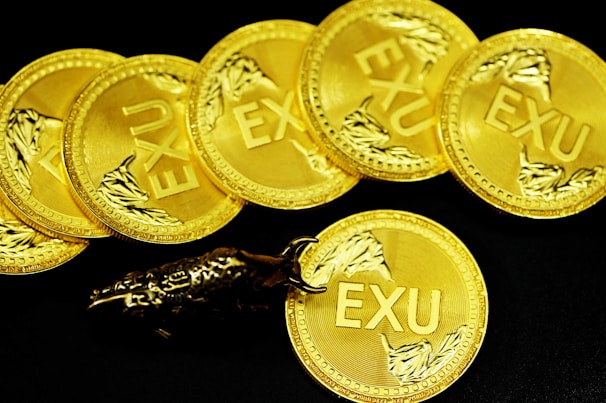 A collection of gold coins with the inscription 'EXU', each decorated with a detailed branch-like design. The coins are laid out in a slightly overlapping arrangement on a black background. A metallic bull figurine rests on one of the coins, adding an element of interest and contrast.