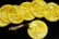 A collection of gold coins with the inscription 'EXU', each decorated with a detailed branch-like design. The coins are laid out in a slightly overlapping arrangement on a black background. A metallic bull figurine rests on one of the coins, adding an element of interest and contrast.