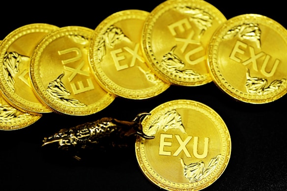 A collection of gold coins with the inscription 'EXU', each decorated with a detailed branch-like design. The coins are laid out in a slightly overlapping arrangement on a black background. A metallic bull figurine rests on one of the coins, adding an element of interest and contrast.