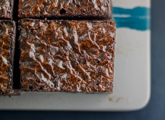 Close-up of a rich chocolate brownie topped with a glossy ganache and delicate gold flakes on a cream-colored plate.