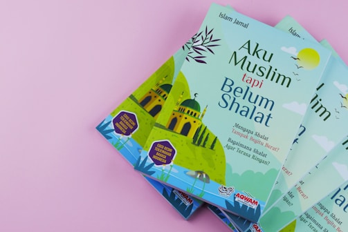 Four copies of a book titled 'Aku Muslim tapi Belum Shalat' are displayed against a pink background. The cover features a mosque illustration with minarets, accompanied by text in Indonesian. The colors are primarily green and blue, with additional accents.