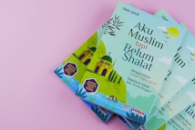 Four copies of a book titled 'Aku Muslim tapi Belum Shalat' are displayed against a pink background. The cover features a mosque illustration with minarets, accompanied by text in Indonesian. The colors are primarily green and blue, with additional accents.
