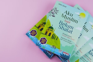 Four copies of a book titled 'Aku Muslim tapi Belum Shalat' are displayed against a pink background. The cover features a mosque illustration with minarets, accompanied by text in Indonesian. The colors are primarily green and blue, with additional accents.
