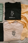 Three t-shirts are neatly arranged on a light-colored surface. The top t-shirt is black and features a graphic of a heart with branches growing from it, accompanied by the text 'Be the love you never received.' A second shirt is pale yellow, showing minimal text, while the third is a light beige color, displaying a detailed heart illustration with the words 'Fear has no place' beneath it. A distinct green item is also partially visible on the left.