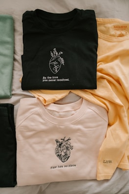 Three t-shirts are neatly arranged on a light-colored surface. The top t-shirt is black and features a graphic of a heart with branches growing from it, accompanied by the text 'Be the love you never received.' A second shirt is pale yellow, showing minimal text, while the third is a light beige color, displaying a detailed heart illustration with the words 'Fear has no place' beneath it. A distinct green item is also partially visible on the left.