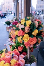 A street-side flower shop displays vibrant bouquets made of orange, yellow, and pink roses. The flowers are arranged in a tree-like structure planted in a pot, surrounded by additional arrangements of roses on the sidewalk. The background shows an urban setting with buildings and blurred traffic.