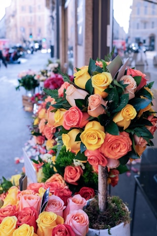 A street-side flower shop displays vibrant bouquets made of orange, yellow, and pink roses. The flowers are arranged in a tree-like structure planted in a pot, surrounded by additional arrangements of roses on the sidewalk. The background shows an urban setting with buildings and blurred traffic.