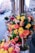 A street-side flower shop displays vibrant bouquets made of orange, yellow, and pink roses. The flowers are arranged in a tree-like structure planted in a pot, surrounded by additional arrangements of roses on the sidewalk. The background shows an urban setting with buildings and blurred traffic.