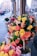 A street-side flower shop displays vibrant bouquets made of orange, yellow, and pink roses. The flowers are arranged in a tree-like structure planted in a pot, surrounded by additional arrangements of roses on the sidewalk. The background shows an urban setting with buildings and blurred traffic.