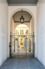 An elegant metal gate with intricate designs stands at the entrance of a covered passageway. The passageway features a high arched ceiling with a hanging lantern-style light fixture. Beyond the gate, the view extends to an exterior courtyard with pale yellow walls and shuttered windows.