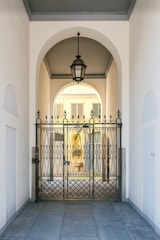 An elegant metal gate with intricate designs stands at the entrance of a covered passageway. The passageway features a high arched ceiling with a hanging lantern-style light fixture. Beyond the gate, the view extends to an exterior courtyard with pale yellow walls and shuttered windows.