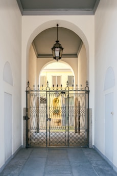 An elegant metal gate with intricate designs stands at the entrance of a covered passageway. The passageway features a high arched ceiling with a hanging lantern-style light fixture. Beyond the gate, the view extends to an exterior courtyard with pale yellow walls and shuttered windows.
