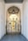An elegant metal gate with intricate designs stands at the entrance of a covered passageway. The passageway features a high arched ceiling with a hanging lantern-style light fixture. Beyond the gate, the view extends to an exterior courtyard with pale yellow walls and shuttered windows.