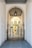 An elegant metal gate with intricate designs stands at the entrance of a covered passageway. The passageway features a high arched ceiling with a hanging lantern-style light fixture. Beyond the gate, the view extends to an exterior courtyard with pale yellow walls and shuttered windows.