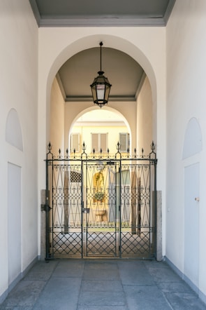 An elegant metal gate with intricate designs stands at the entrance of a covered passageway. The passageway features a high arched ceiling with a hanging lantern-style light fixture. Beyond the gate, the view extends to an exterior courtyard with pale yellow walls and shuttered windows.