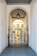 An elegant metal gate with intricate designs stands at the entrance of a covered passageway. The passageway features a high arched ceiling with a hanging lantern-style light fixture. Beyond the gate, the view extends to an exterior courtyard with pale yellow walls and shuttered windows.
