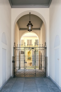 An elegant metal gate with intricate designs stands at the entrance of a covered passageway. The passageway features a high arched ceiling with a hanging lantern-style light fixture. Beyond the gate, the view extends to an exterior courtyard with pale yellow walls and shuttered windows.
