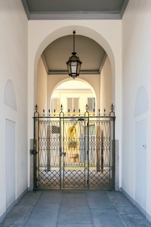 An elegant metal gate with intricate designs stands at the entrance of a covered passageway. The passageway features a high arched ceiling with a hanging lantern-style light fixture. Beyond the gate, the view extends to an exterior courtyard with pale yellow walls and shuttered windows.