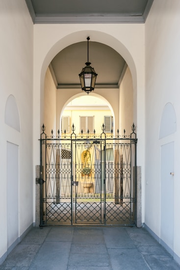 An elegant metal gate with intricate designs stands at the entrance of a covered passageway. The passageway features a high arched ceiling with a hanging lantern-style light fixture. Beyond the gate, the view extends to an exterior courtyard with pale yellow walls and shuttered windows.