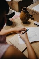 A person is sketching on a piece of paper placed on a desk. The sketch resembles an architectural drawing or design. On the desk, there are several objects including a rustic vase, a small black pedestal, a ruler, and a book. There are also cardboard boxes in the background with some writing on them.
