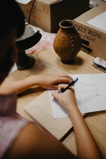 A person is sketching on a piece of paper placed on a desk. The sketch resembles an architectural drawing or design. On the desk, there are several objects including a rustic vase, a small black pedestal, a ruler, and a book. There are also cardboard boxes in the background with some writing on them.