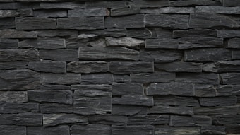 A wall constructed from tightly stacked, rectangular slate stones. The stones vary slightly in size and texture, creating a rugged and natural appearance. The overall color is a dark gray, with subtle variations throughout the surface.