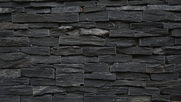 A wall constructed from tightly stacked, rectangular slate stones. The stones vary slightly in size and texture, creating a rugged and natural appearance. The overall color is a dark gray, with subtle variations throughout the surface.