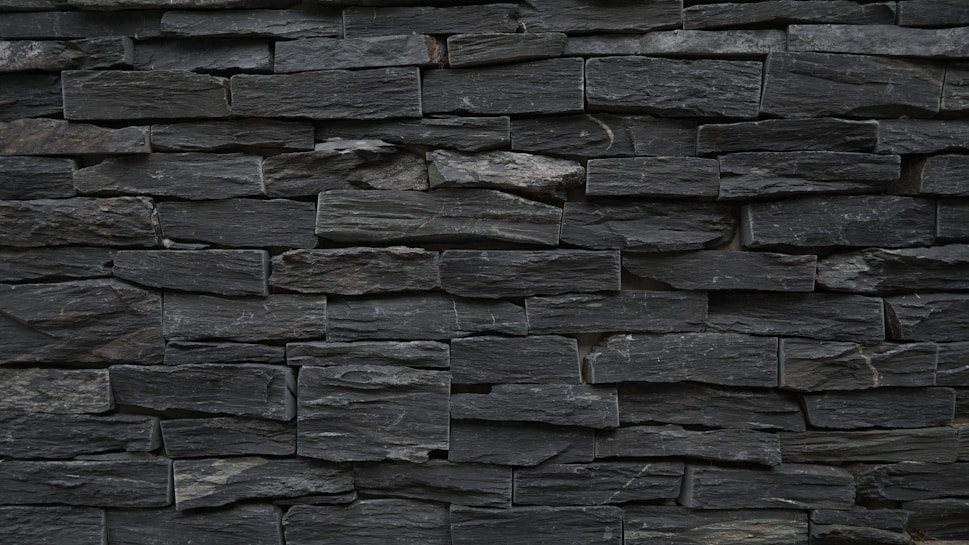 A wall constructed from tightly stacked, rectangular slate stones. The stones vary slightly in size and texture, creating a rugged and natural appearance. The overall color is a dark gray, with subtle variations throughout the surface.