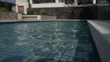 A modern swimming pool with clear water reflecting the light, surrounded by stone and concrete walls. A building with large windows is visible in the background, along with some greenery.