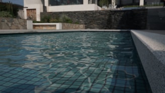 A modern swimming pool with clear water reflecting the light, surrounded by stone and concrete walls. A building with large windows is visible in the background, along with some greenery.