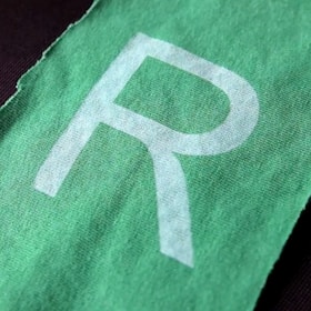 A close-up view of a green fabric with a large white letter 'R' printed on it. The fabric is textured, and the edges appear slightly frayed.