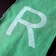 A close-up view of a green fabric with a large white letter 'R' printed on it. The fabric is textured, and the edges appear slightly frayed.