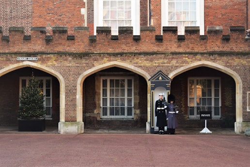 Two uniformed guards are standing in front of an arched doorway of a brick building with a fortress-like facade. There is a Christmas tree in a pot on the left, adjacent to one of the arches, and a 'PRIVATE' sign on the right. The area is a clean, paved courtyard.