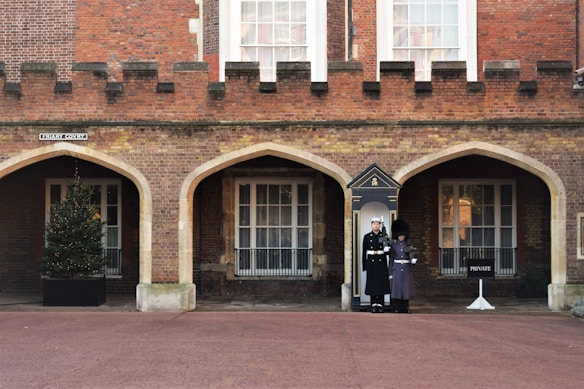 Two uniformed guards are standing in front of an arched doorway of a brick building with a fortress-like facade. There is a Christmas tree in a pot on the left, adjacent to one of the arches, and a 'PRIVATE' sign on the right. The area is a clean, paved courtyard.
