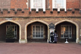 Two uniformed guards are standing in front of an arched doorway of a brick building with a fortress-like facade. There is a Christmas tree in a pot on the left, adjacent to one of the arches, and a 'PRIVATE' sign on the right. The area is a clean, paved courtyard.