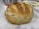 A round, rustic loaf of bread with a golden-brown crust and visible seeds or grains baked into the surface. It rests on a sheet of parchment paper on a countertop, with a metal container and cloth in the background.