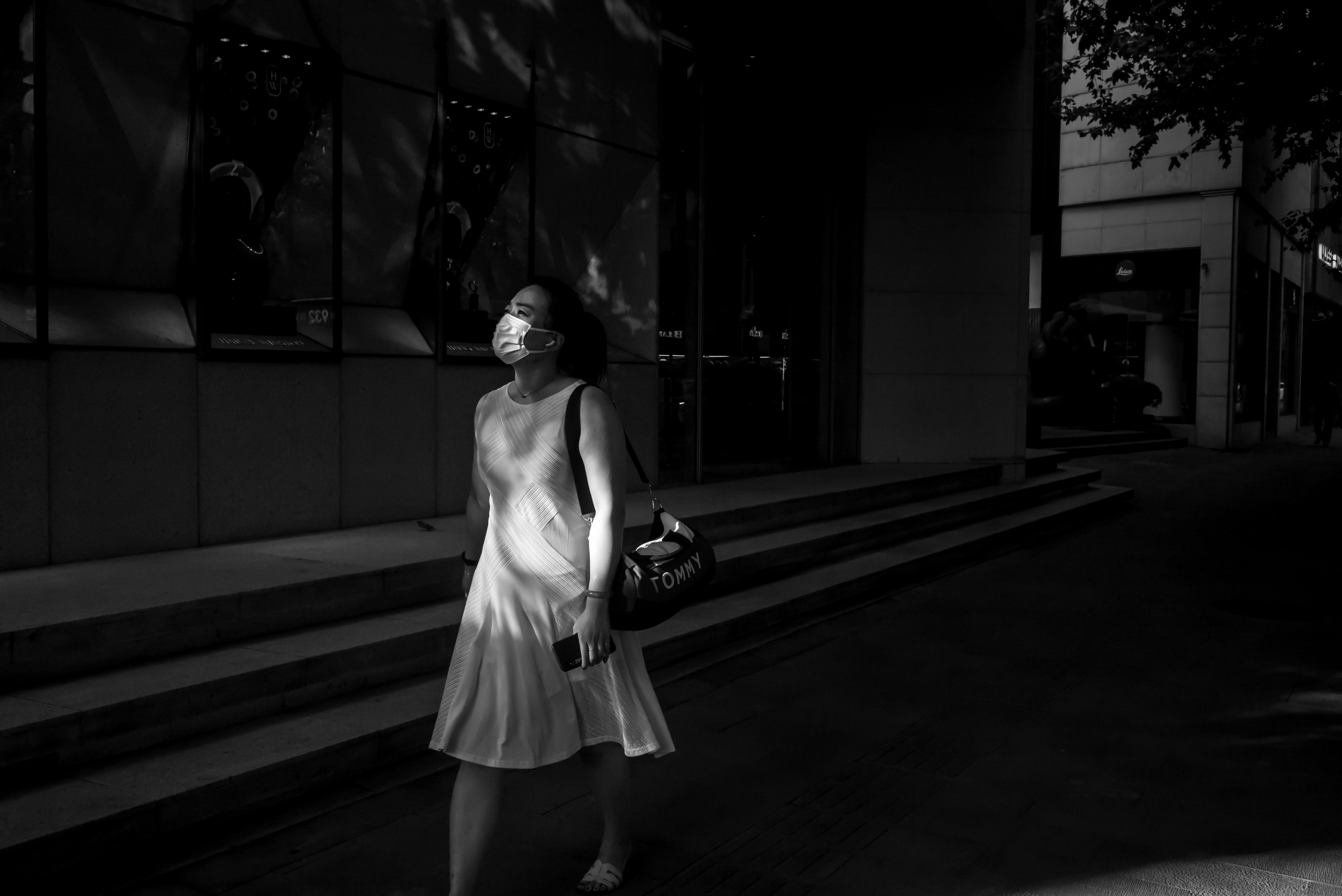 Black-and-white photograph of a woman in a white dress and mask walking down steps beside glass storefronts.