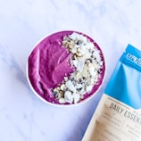 A bowl filled with vibrant purple smoothie, topped with a variety of nuts and seeds, is placed on a light surface. To the right of the bowl, a package labeled 'Daily Essentials' in blue and white coloring is visible.