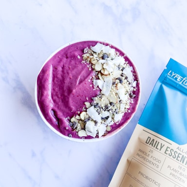 A bowl filled with vibrant purple smoothie, topped with a variety of nuts and seeds, is placed on a light surface. To the right of the bowl, a package labeled 'Daily Essentials' in blue and white coloring is visible.