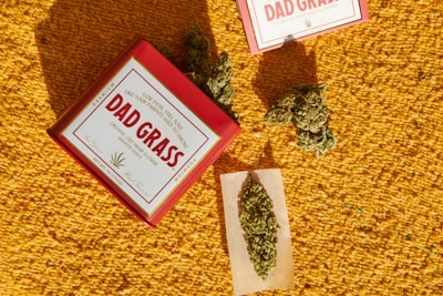 A vibrant yellow textured background sets the scene for a reddish box labeled 'Dad Grass', which is partially open with some dried green plant material visible. Nearby, more plant materials and a rolling paper with some of the same substance are positioned.