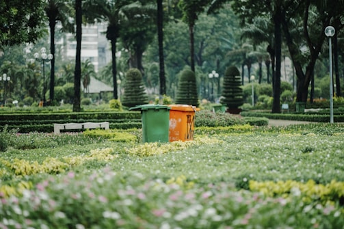A lush green park featuring well-manicured hedges and a variety of trees, with two colorful trash bins located prominently at the center. Pathways and benches are also visible, surrounded by dense foliage.