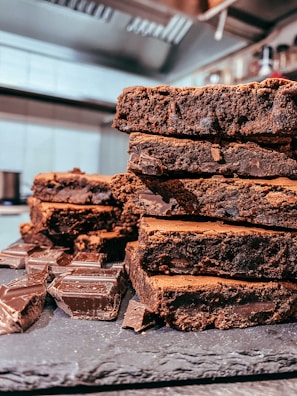A stack of rich, chocolatey brownies is placed on a slate board, with chunks of dark chocolate scattered around. The brownies have a dense, moist texture, and the background includes a blurred view of a kitchen setting.