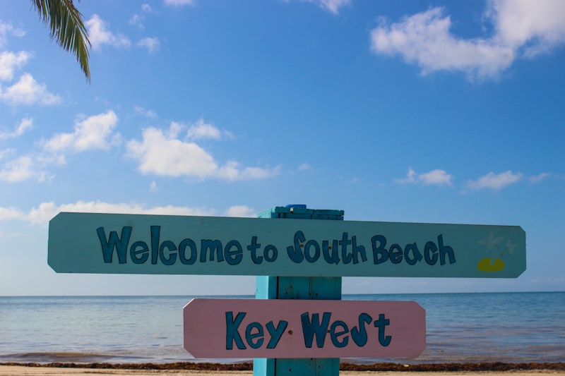 Hand-painted wooden signs reading Welcome to South Beach and Key West mounted on a post against a blue sky and calm sea