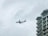 An airplane is in flight against a cloudy sky, with a multi-story building on the right side of the image. The building has balconies and a distinct architectural design on the roof.
